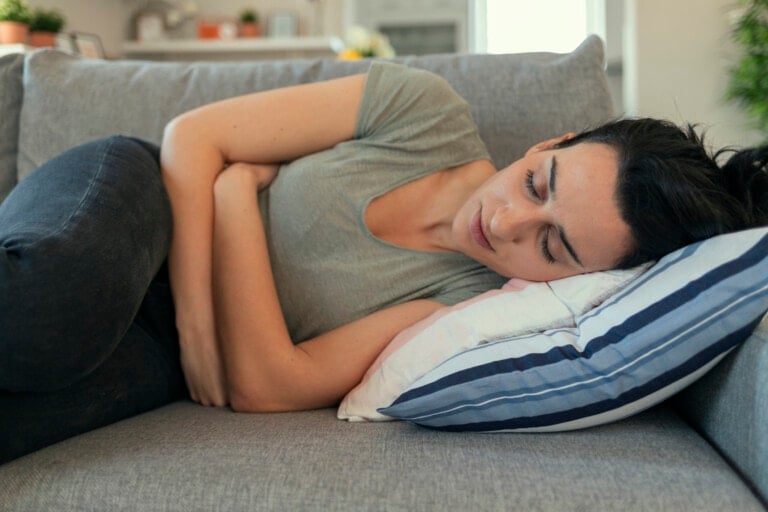 A woman with dark hair, wearing a green shirt and black pants, lies on her side on a gray couch. Resting her head on a white pillow with blue stripes, she has her arms folded across her stomach, perhaps experiencing pregnancy symptoms. The background includes a blurred kitchen setting.