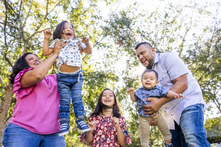 A family of five is outdoors on a sunny day with trees in the background. The mother in a pink shirt is lifting a young girl, embracing body positivity, while the father in a light blue shirt is holding a baby. An older girl stands in the middle, smiling confidently.
