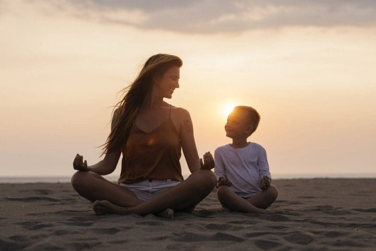 A woman and a child are sitting cross-legged on a beach at sunset, facing each other and smiling. Both have their hands resting on their knees in a meditative pose, deeply immersed in meditation. The sky is partly cloudy with a warm, golden hue from the setting sun.