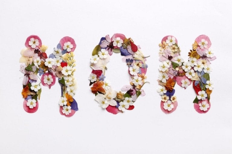 The word "MOM" is spelled out using a variety of colorful flowers and petals arranged on a white background. The letters are formed by an assortment of blooms, including white, pink, purple, and yellow flowers—perfect Mother's Day gifts for pregnant mamas celebrating their journey.