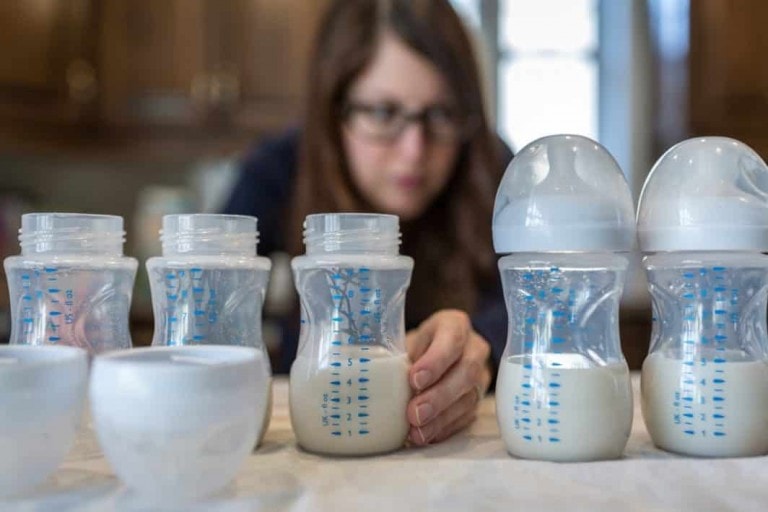 A person with glasses, slightly out of focus, is preparing formula for baby by placing baby bottles filled with milk on a countertop. Five baby bottles, with measurement markings and capped with white lids, are aligned in the foreground of the image.