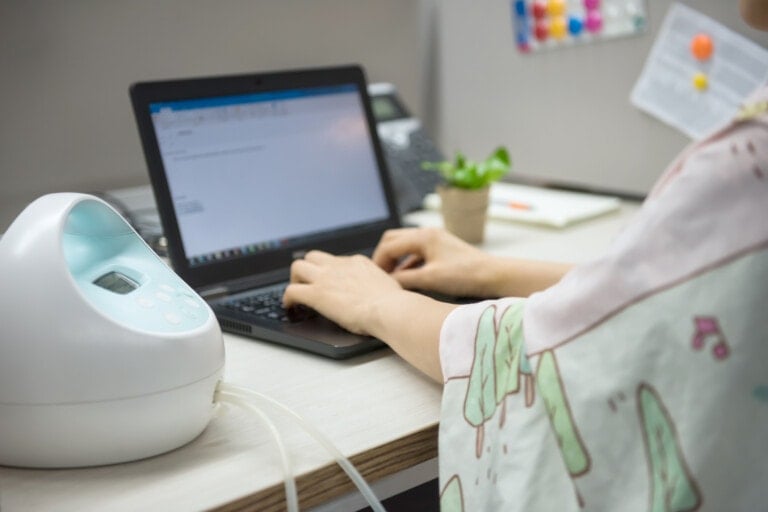 A person is typing on a laptop at a desk. Next to them is a breast pump machine, hinting at tips for pumping at work. A small potted plant and some stationery items are also on the desk. The background shows a gray wall with a colorful bulletin board.