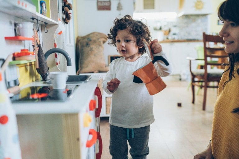 A young child with curly hair wearing a white shirt and gray pants stands in front of one of the best play kitchens for toddlers, holding an orange kettle. An adult, partially visible, watches from the side. The kitchen features various toy utensils and dishes.
