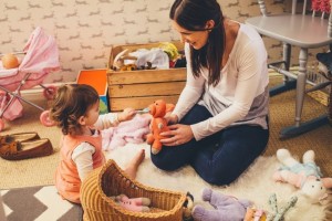 A toddler and an adult sit on a carpeted floor in a playroom. The toddler, wearing an orange dress, plays with a stuffed bear, while the occupational therapist, wearing a white long-sleeve shirt and blue jeans, watches and interacts. Toys and a wicker basket are scattered around them.