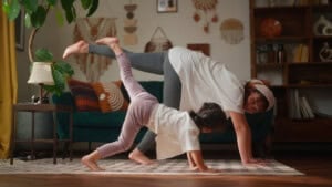An adult and a child practice yoga together indoors, both in a downward dog pose with one leg raised. The room is decorated with plants, wall art, and a green sofa.