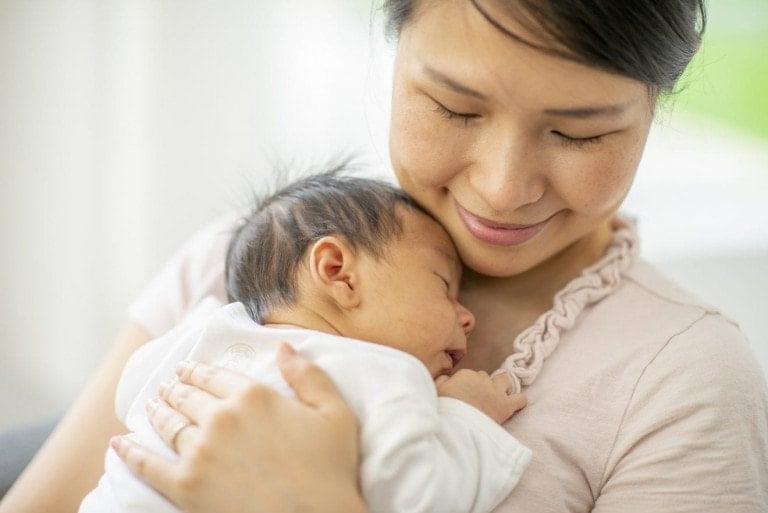 A woman holds a sleeping baby against her chest, smiling softly in a light pink top. The baby, dressed in a white outfit, looks peaceful and content. The scene exudes calm and nurturing warmth—perfect inspiration for baby gifts for newborns.