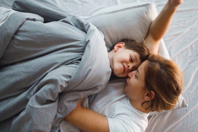 A woman and a young boy lie in bed under a grey blanket. The woman, with light brown hair, kisses the boy on the cheek as he smiles. The boy has dark hair, and the bedsheet is light grey. They seem to be sharing an affectionate moment typical when kids sleep in bed with me.