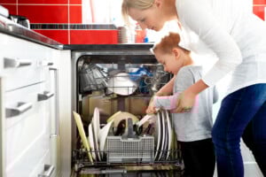 An adult and a child are learning valuable life skills as they load dishes into an open dishwasher in a brightly lit kitchen. The countertop is black, and the backsplash tiles are red. The dishwasher contains various dishes, including plates, bowls, and cutlery.