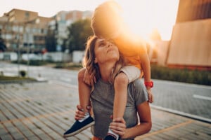 A woman is carrying her firstborn on her shoulders outdoors on a sunny day. The child is wearing a red wristband and blue shoes, while the woman is in a gray shirt. They are sharing a moment of affection with a sunset and buildings in the background.