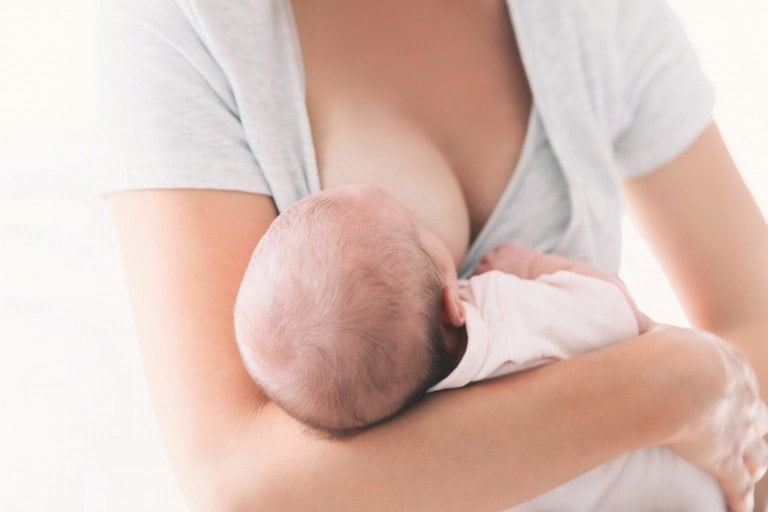 A woman is breastfeeding a baby in the cross cradle hold. The baby's head is cradled in her arm, latching onto the breast. She is wearing a light gray top while the infant wears a white outfit. The background is out of focus, emphasizing this intimate moment.