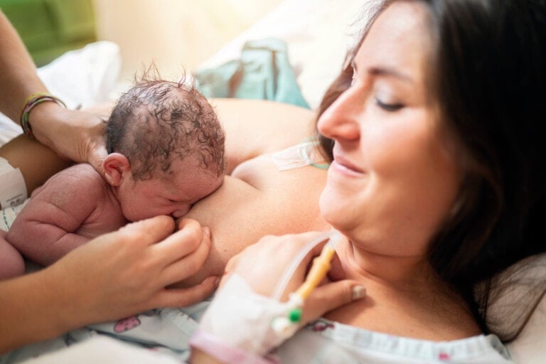 A newborn baby with wet hair is pressed against its mother's chest, breastfeeding for the first time after a c-section. The mother, wearing a hospital gown and with an IV in her hand, is lying on a bed, smiling gently and looking at the baby.
