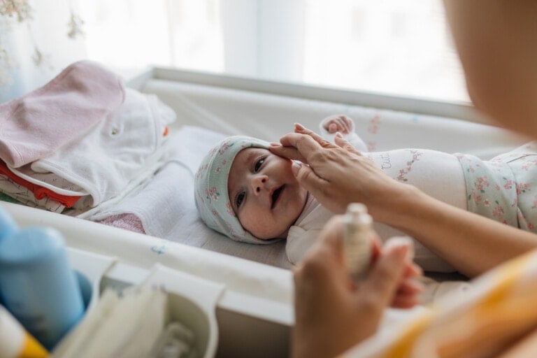 A baby lies on a changing table wearing a floral-patterned hat and onesie, while an adult gently touches the baby's face, ensuring the baby's skin is protected. Nearby are folded clothes and baby supplies. The background is softly lit by natural light from a window.