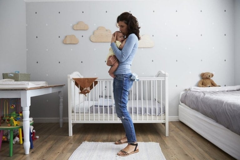 A woman stands in a nursery holding a baby, gently rocking to alleviate the baby's reflux. The room features a white crib, changing table, bed, and various toys. The walls are adorned with cloud and drop-shaped wall stickers. A stuffed teddy bear sits on the bed in the background.
