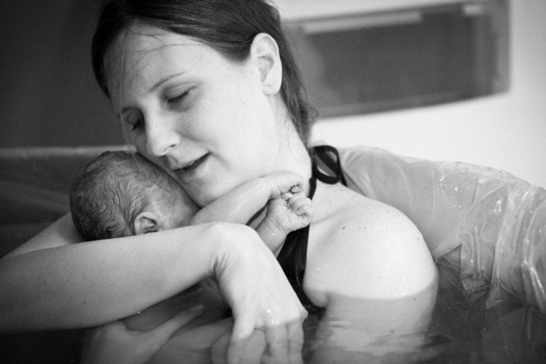 A woman in a water birthing pool holds a newborn baby in her arms, embodying the serene beauty often discussed on home birth podcasts. She has her eyes closed and is smiling gently. The baby rests against her chest with its tiny hands and feet visible. The image is in black and white.