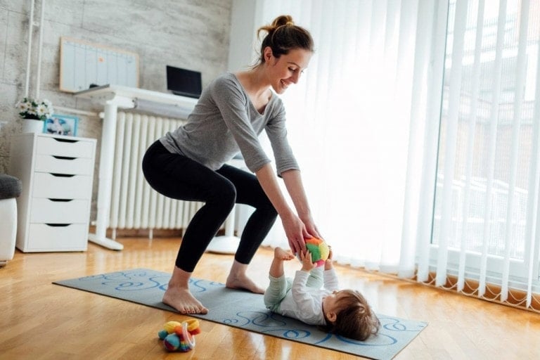 A woman is doing squats while holding a baby who is lying on a yoga mat, both smiling. There is a stuffed toy on the floor next to the mat. The room has white, vertical blinds and a desk in the background, showing how you can effortlessly include your kids in your workout routine.