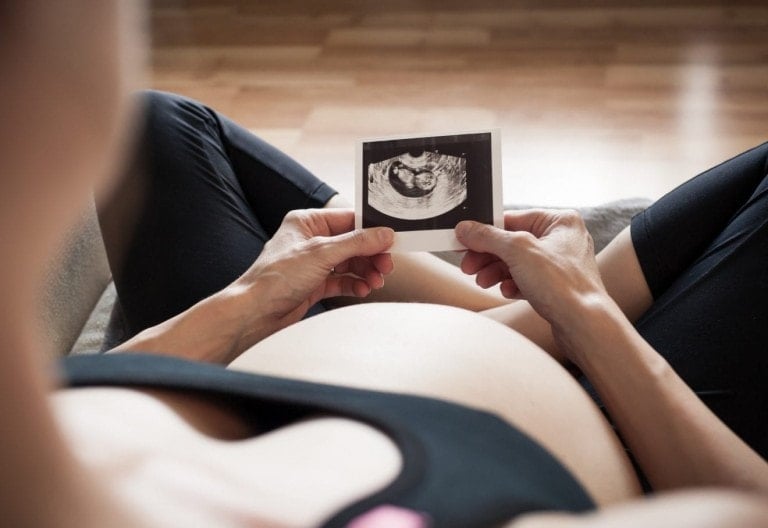 A person with a pregnant belly is sitting on the wooden floor, holding an ultrasound image in their hands. They're dressed in a black top and pants, looking serene as they listen to a pregnancy affirmations podcast.