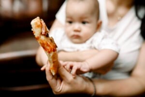 A baby, held by a person in a white shirt, looks intently at a slice of pizza with toppings, which is being held close to it by another hand. The baby appears curious and reaches out toward the pizza, showing an early interest in first foods. The background is blurred.