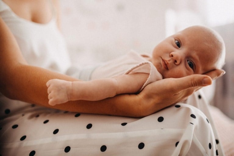A baby lies contently on an adult's lap, supported by the adult's hand. The baby, dressed in light-colored clothing, gazes up with curiosity, maybe a future in STEM learning awaits. The adult is wearing a white dress with black polka dots, and the background appears soft and out of focus.