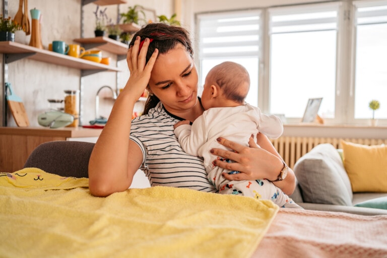 A woman sits in a living room holding a baby while resting her head on her hand, possibly experiencing postpartum hormone changes. The baby, facing the woman and dressed in a white shirt with colorful patterned pants, is surrounded by calmness. A yellow towel is spread on a table in front of them, with a couch visible in the background.