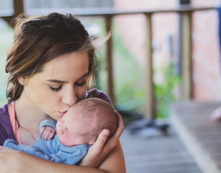 A woman holds a sleeping newborn wrapped in a blue blanket and kisses the baby on the forehead, trusting her instincts. The setting appears to be a porch with blurred background elements.