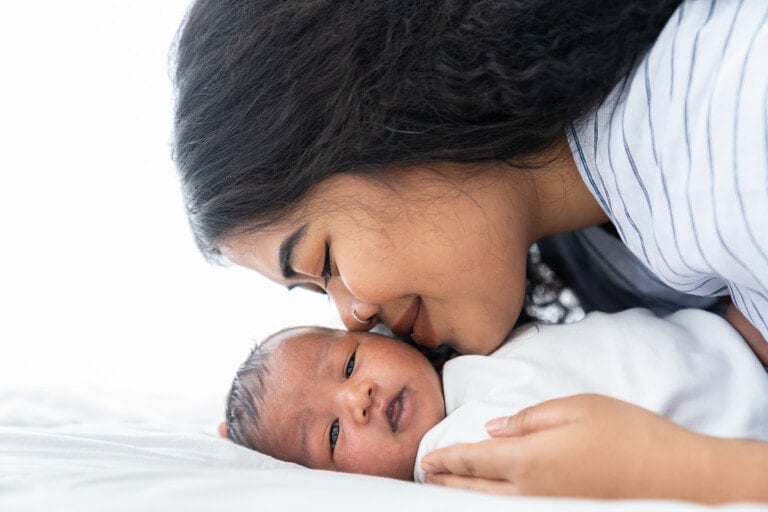 A new mom with long dark hair is lying next to a newborn baby who is wearing a white outfit. She leans in to kiss the baby's forehead. Both are lying on a white surface, with the baby’s eyes open, looking slightly towards the camera.