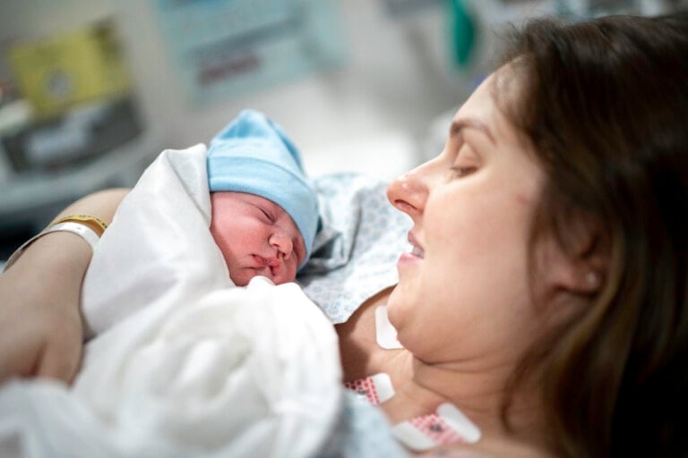 A woman lies in a hospital bed, holding a newborn baby wrapped in a white blanket. The baby, wearing a blue cap, appears to be sleeping. The woman, experiencing postpartum shaking, has her eyes closed and is smiling. Medical equipment and supplies can be seen blurred in the background.