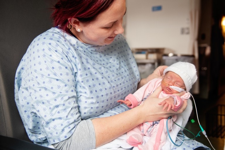 A woman wearing a hospital gown holds a premature baby in her arms. The baby, dressed in pink clothing and a white hat, is connected to medical tubes. They are in a medical setting with blurred equipment in the background, highlighting the need to help a NICU mom during this challenging time.