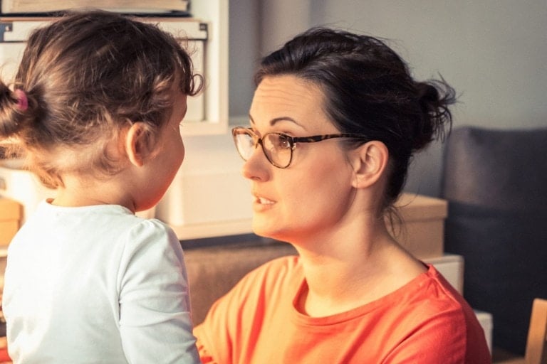 A woman wearing glasses and a red top faces and talks to a young child with curly hair, who is wearing a light-colored shirt. They are indoors, surrounded by shelves and storage boxes. This moment captures an invaluable parenting hack as she elegantly imparts wisdom to the attentive child.