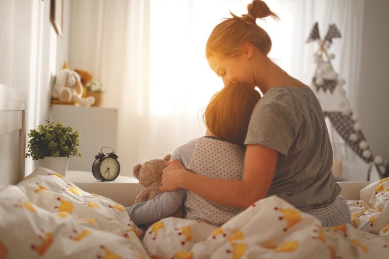 A woman with excellent parenting skills sits on a bed hugging a child who is holding a teddy bear. The room is softly lit by sunlight and decorated with a plant, an alarm clock, and stuffed toys. The bedding has a duck pattern.