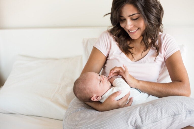 A woman sits on a bed, smiling down at a baby she is holding. The baby is wrapped in a blanket and lying on a cushion on her lap, which doubles as a nursing pillow. The background consists of white bedding and pillows.
