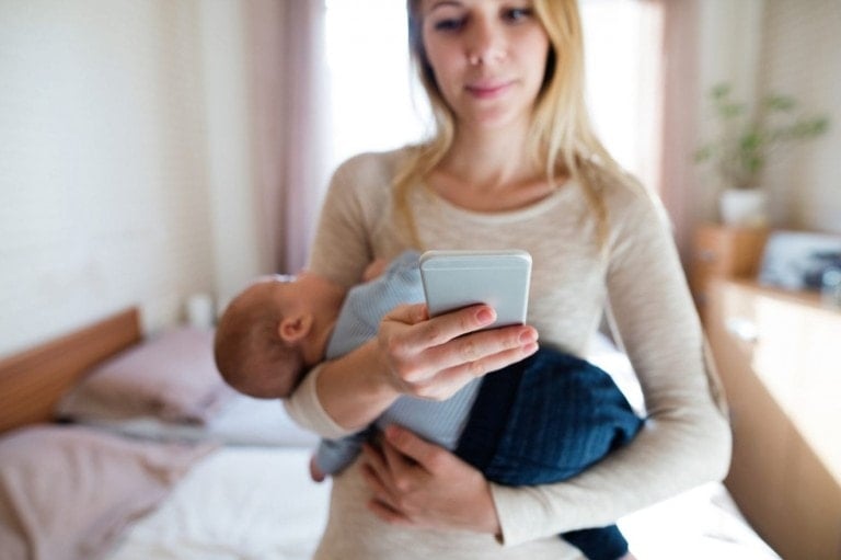 A woman, likely a new parent, holds an infant in her arms while looking at baby apps on her smartphone. The setting appears to be a bedroom with a bed in the background. The woman is wearing a beige long-sleeved shirt, and the baby is dressed in a blue outfit.