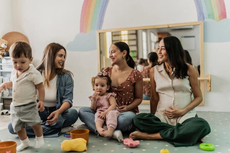 Three women sit on the floor of a playroom, smiling and sharing opinions. Two young children are also present; one is standing and walking while the other sits on a woman's lap. The room features child-friendly decor with a rainbow mural and various toys scattered on the floor.