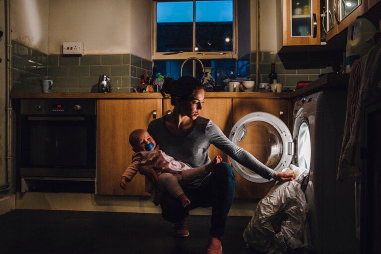A woman is kneeling on the floor of a dimly lit kitchen, holding a baby while loading clothes into a front-loading washing machine. The light from the machine softly illuminates her and the baby, capturing a quiet moment of motherhood. The kitchen features wooden cabinets and a tiled backsplash.