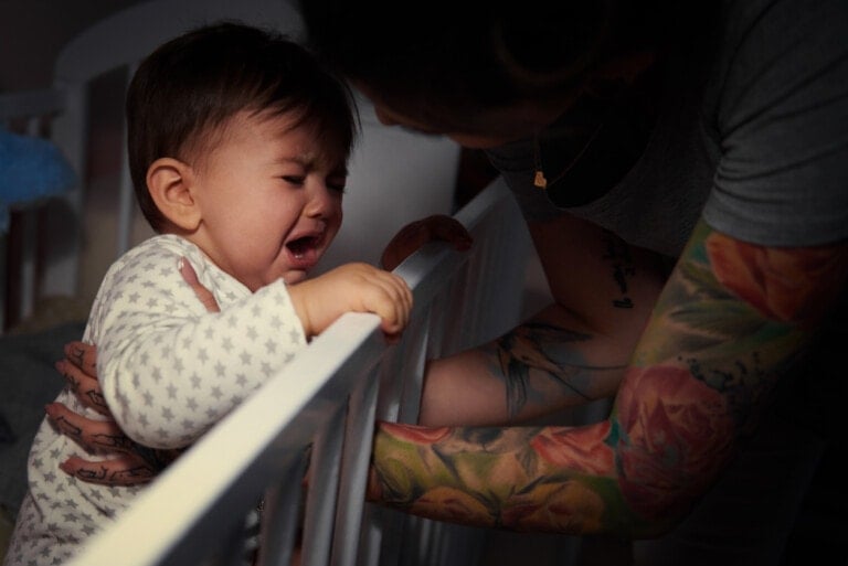 A baby is crying while standing in a crib, holding the rail. An adult with tattooed arms reaches into the crib, possibly comforting the baby or helping out. The dimly lit room suggests a quiet moment to consider why your baby wakes up crying.