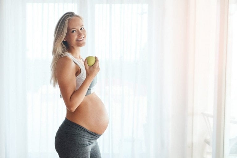 A pregnant woman stands sideways, smiling at the camera. She is wearing a white sports bra and gray leggings, holding a green apple in her right hand—a perfect snack to meet her body needs during pregnancy. The background features light-colored curtains with natural light streaming through the windows.
