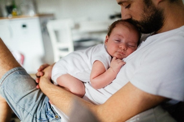 A man sitting and cradling a sleeping baby on his chest in a cozy indoor setting. The baby, dressed in a white onesie, is blissfully napping while the dad, wearing a white t-shirt and denim shorts, relishes this tender moment. It's heartwarming to see dad baby bonding like this.