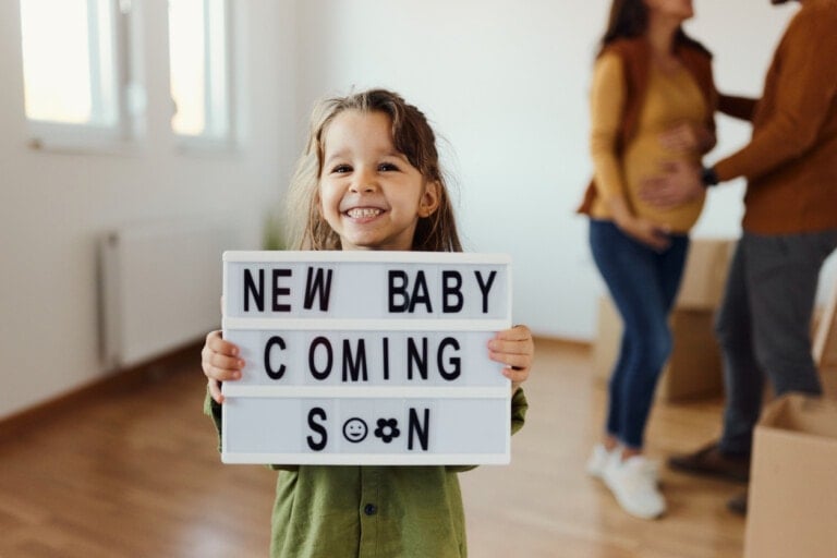 A smiling child holds a sign that reads New Baby Coming Soon in a brightly lit room. In the background, a woman with a baby bump and a man are standing together, slightly out of focus, depicting the perfect moment when to announce pregnancy.