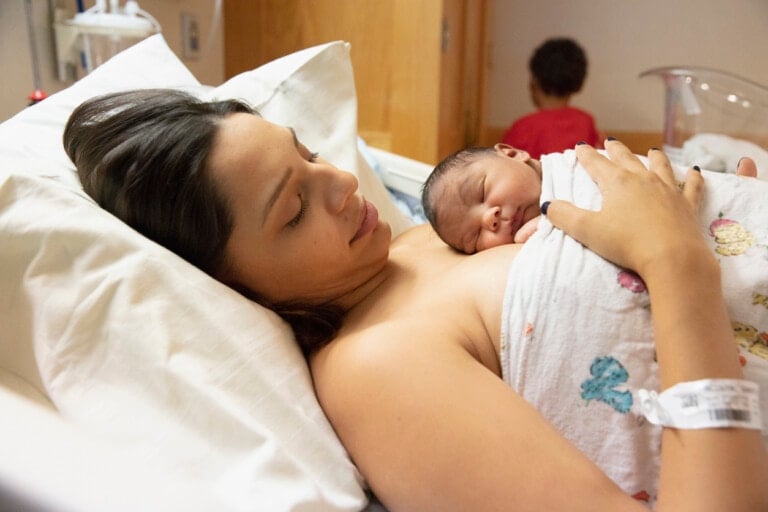 A woman rests in a hospital bed holding a newborn baby wrapped in a colorful blanket on her chest. She wears an identification bracelet on her wrist, having just overcome postpartum hemorrhage. In the background, a child in a red shirt faces away from the camera.
