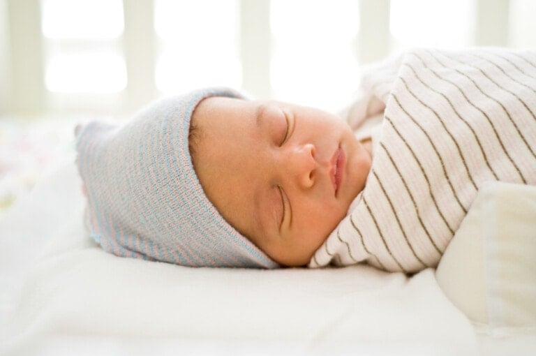 A newborn baby is peacefully sleeping, swaddled in one of the best swaddles—a white blanket with gray stripes. The baby is wearing a light blue knitted hat. The background is softly lit, creating a calm and serene atmosphere.