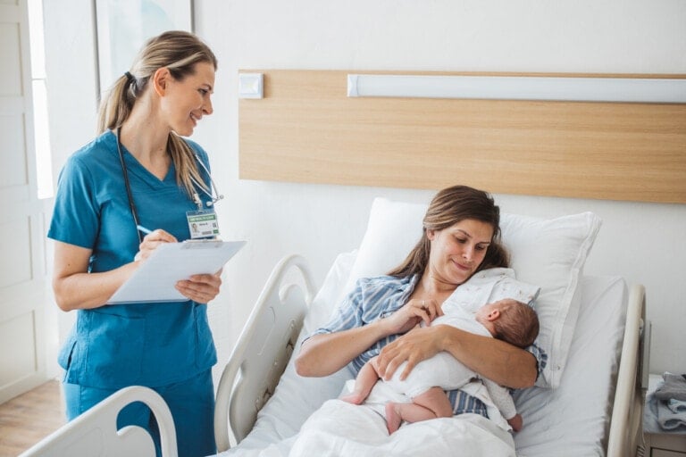 A postpartum nurse in blue scrubs is holding a clipboard and speaking to a woman lying in a hospital bed, who is cradling and bottle-feeding a baby. The hospital room has white walls and a wooden headboard.