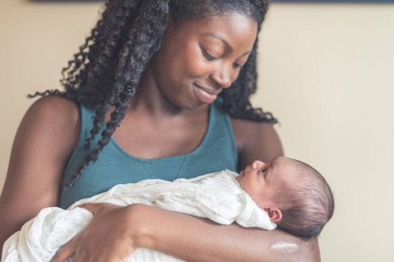 A woman with long curly hair, in the midst of gathering her c-section recovery must-haves, is holding a newborn baby wrapped in a light blanket. She is looking down and smiling at the baby while standing indoors.