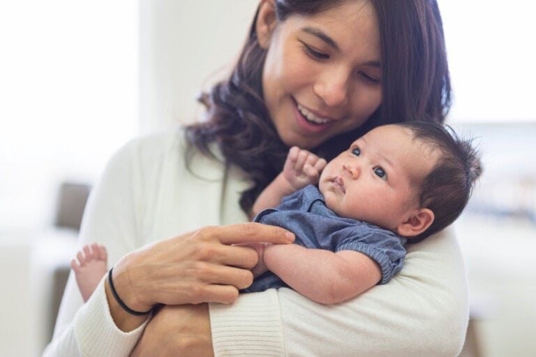 A first-time mom is smiling and holding a baby dressed in a blue outfit. The woman is looking at the baby, who appears to be gazing off to the side. They are indoors with a blurred background.