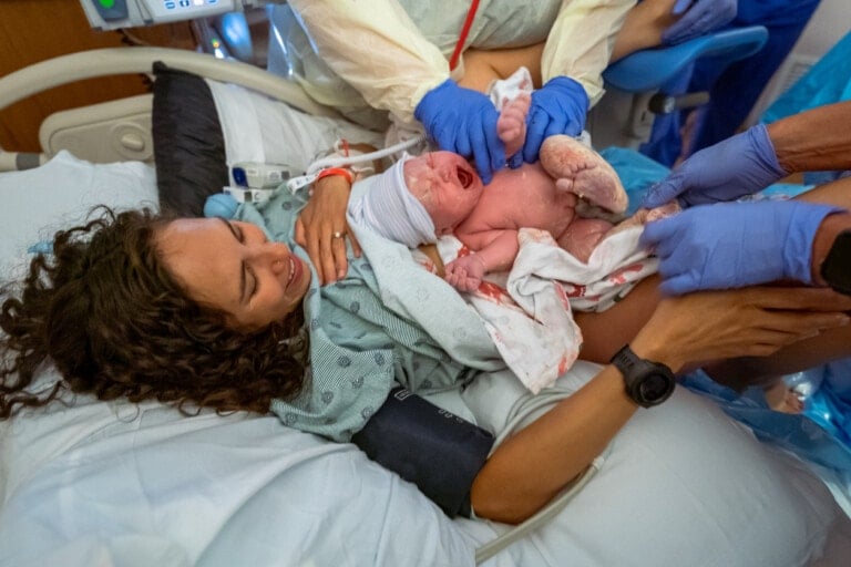 A newborn baby is being held by medical staff wearing gloves and hospital attire. The baby, possibly responding to the effects of pitocin administered during delivery, is crying and appears to be freshly delivered. The mother, lying in a hospital bed, has her arm around the baby. Medical equipment is visible in the background.