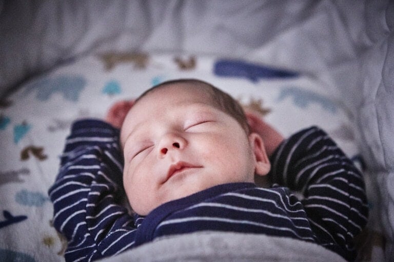 A friend's newborn is peacefully sleeping with their arms raised above their head. The baby, who sleeps through the night, is wearing a dark striped shirt and lying on a patterned blanket adorned with animals and shapes. The scene appears serene and cozy.