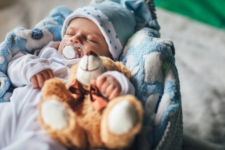 A baby boy wearing a white outfit and a blue hat with star patterns sleeps in a blue and white blanket. The baby has a pacifier in their mouth and is holding a brown stuffed bear, bringing joy to anyone who've longed to conceive a boy.