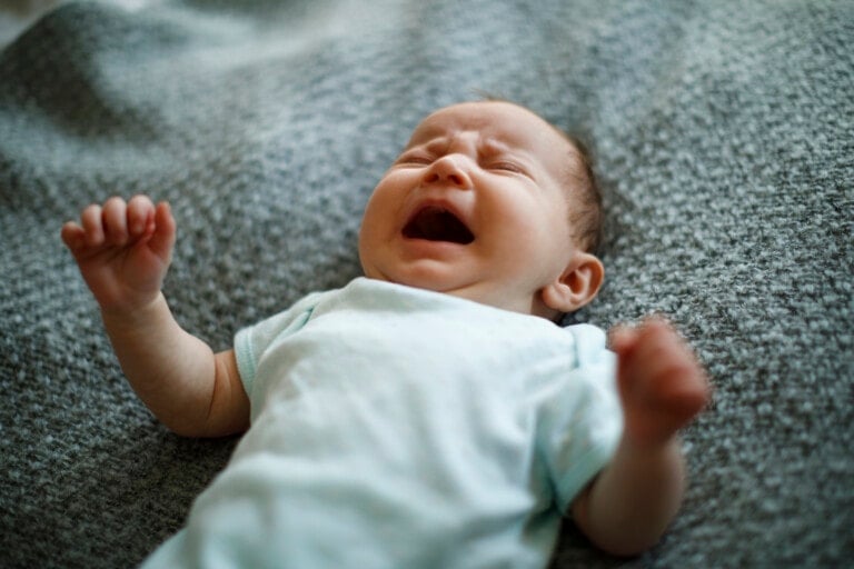 A baby wearing a light blue onesie is lying on a textured grey blanket. With both arms raised and appearing to cry or wail, the scene reflects normal baby fussiness, suggesting the little one may be upset or uncomfortable.