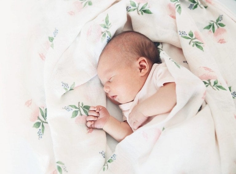 A sleeping baby is swaddled in a white blanket with a floral pattern, much like the serene scenes depicted in baby sleep books. The infant, with a light complexion and light-colored outfit, rests peacefully within the blanket adorned with green leaves and pink flowers.