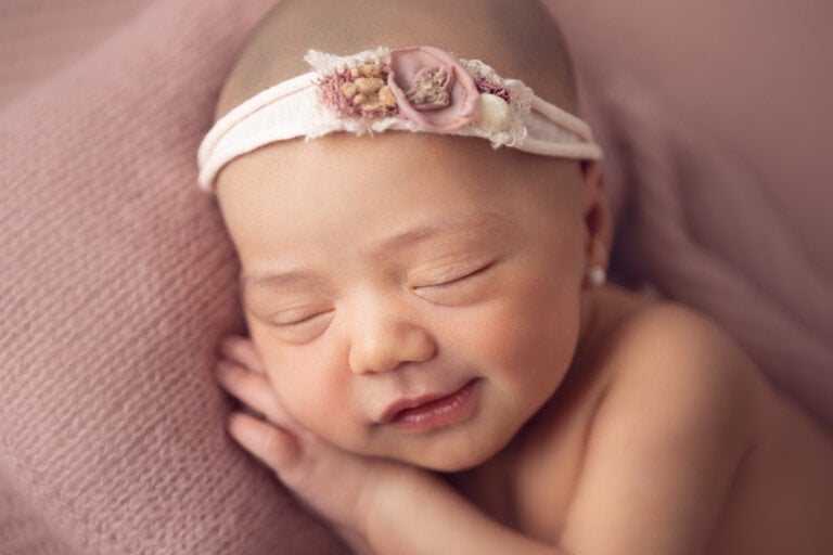 A newborn baby sleeps peacefully, resting its head on a soft, pink cushion. The baby, perhaps destined for one of those cute baby girl names, is wearing a white headband with floral decorations and has a serene smile on its face. The background is softly blurred in shades of pink.