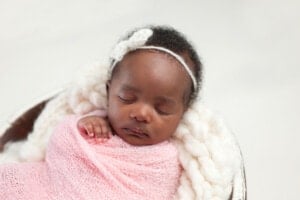 A sleeping baby wrapped in a pink blanket is lying in a cushioned basket. The baby, possibly one of those adorable girl names with E, is wearing a white knitted headband with a bow. The background is soft and out of focus, emphasizing the peaceful scene.