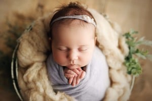 A baby girl, potentially with names like Bella or Brianna, is sleeping peacefully with hands folded under the chin, wrapped in a soft gray blanket, and wearing a headband with a tiny flower. The baby is nestled in a fluffy off-white cushion with some green leaves around it.
